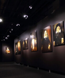 Una galleria d'arte scarsamente illuminata espone una fila di ritratti classici, appesi a pareti scure e illuminati dai faretti PC LED 100W Serie Hyperion Teatri e Studi TV. I dipinti raffigurano vari individui in abiti formali, creando un'atmosfera tranquilla e riflessiva nella stanza.