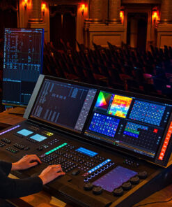 Una persona aziona una console di controllo luci Eos Apex 10, che presenta più schermi che mostrano varie impostazioni e schemi di colori. La console è installata in un auditorium, con file di sedili vuote visibili sullo sfondo.