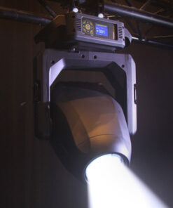 Un riflettore da palco Lonestar con un involucro nero è appeso a una capriata metallica, emettendo un fascio di luce bianca brillante. La luce è accesa, illuminando l'area sottostante, con un piccolo display digitale e un pannello di controllo visibili in alto.