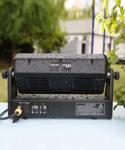 Una luce da palcoscenico per esterni Sirena, di colore nero, è posizionata su una superficie azzurra adornata da gocce d'acqua. È circondata dal verde in un ambiente da giardino, a suggerire una recente pioggia.