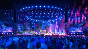 Una scena di concerto vibrante in un anfiteatro all'aperto, con una grande folla di fronte a un palco illuminato. Il palco è adornato con luci blu e rosa, creando un'atmosfera energica. Gli artisti sono visibili, insieme a grandi schermi su ogni lato.